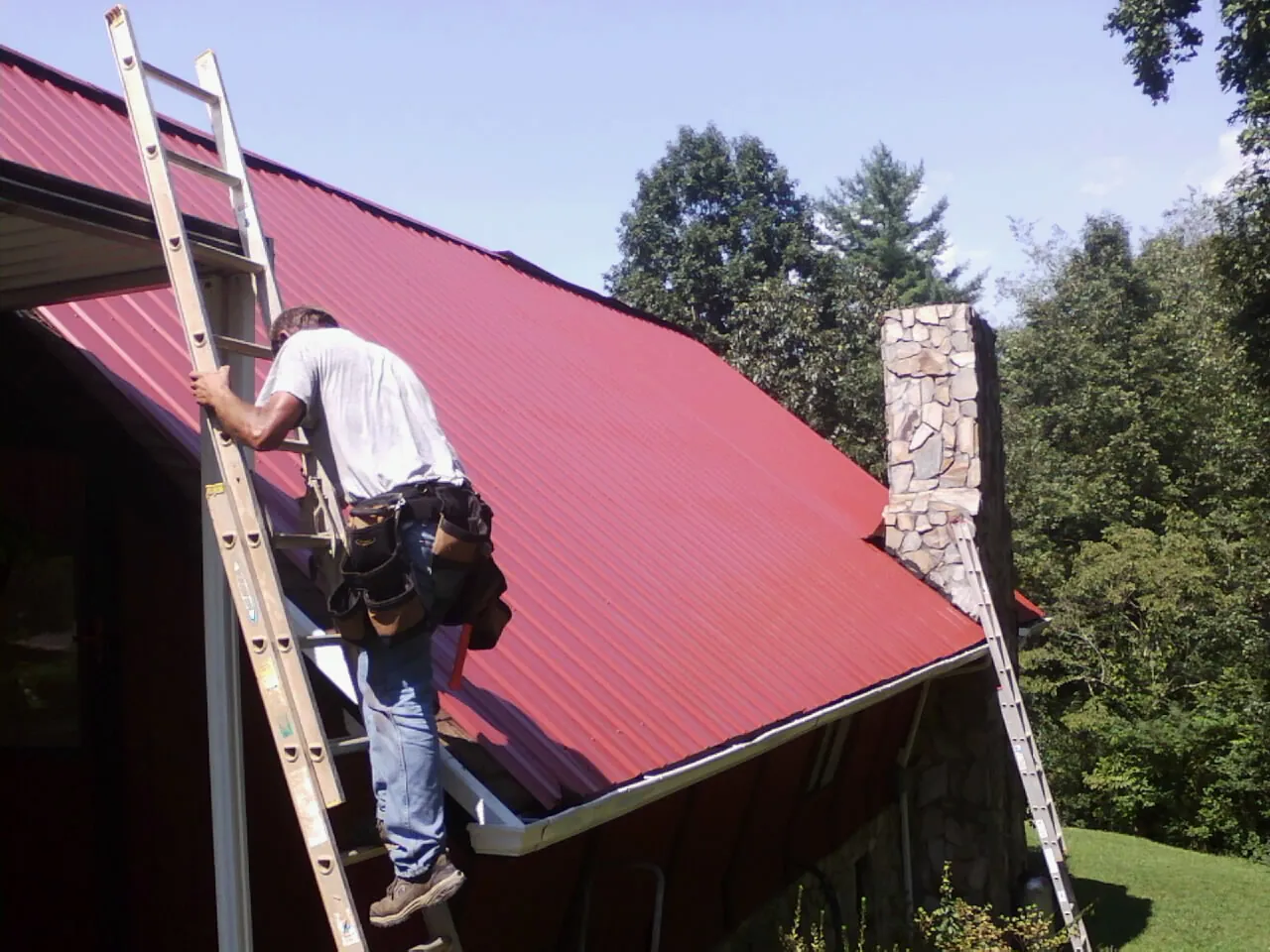 Roofer on a ladder beside a red metal roof and stone chimney — Soffit & Fascia Repair by Roofing Brothers in Woodbourne